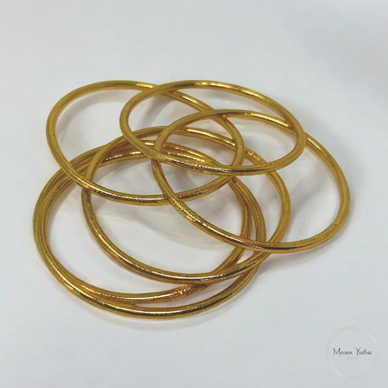 May include: A collection of gold-colored bangles, intertwined and arranged on a white surface. The bangles are circular and appear to be made of metal. The image is well-lit, highlighting the metallic sheen of the jewelry.