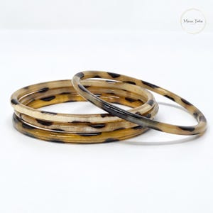 Op de afbeelding: Een set van drie ronde armbanden met een doorschijnend beige en bruin schildpadpatroon. De armbanden hebben een gladde, gepolijste afwerking met donkerbruine vlekken verdeeld over de lichtbruine basis, wat zorgt voor een gelaagd effect.