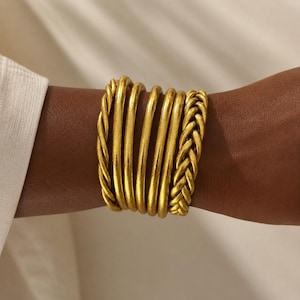 Peut inclure: Un bracelet manchette doré au design multi-brins. La pièce se compose de plusieurs bandes de fil arrondies et lisses disposées côte à côte, avec une seule bande à texture tressée sur le bord droit, créant un aspect large et superposé.
