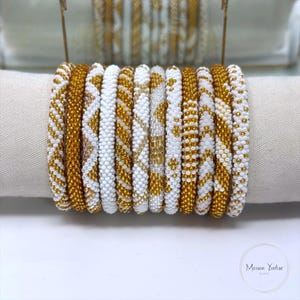 Op de afbeelding: Een set van elf cilindrische armbanden met gedetailleerd kraalwerk in wit en metallic goud. De armbanden tonen diverse patronen zoals diagonale strepen, chevron zigzaglijnen en effen banden, allemaal gemaakt van kleine, uniforme glaskralen.