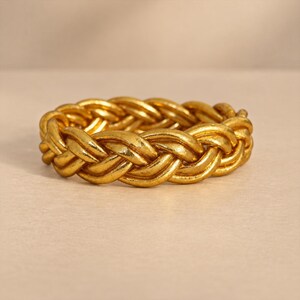 Peut inclure: Un bracelet doré présentant un motif tressé continu. La surface métallique possède une finition polie et réfléchissante avec une texture subtile sur l'ensemble des brins entrelacés, formant un anneau circulaire à l'aspect métallique uniforme.