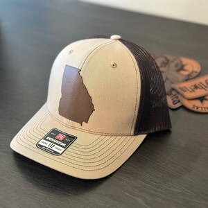 May include: A tan and brown trucker hat with a brown leather patch of the state of Georgia on the front. The hat is made by Richardson and has a black mesh back.