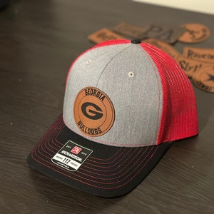 May include: A gray and red trucker hat with a black bill. The hat has a brown leather patch with the text "GEORGIA BULLDOGS" and a large "G" in the center.