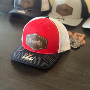 May include: Red, white, and navy blue baseball cap with a brown leather patch that says "Braves" in white script. The patch also features a baseball bat and an axe.