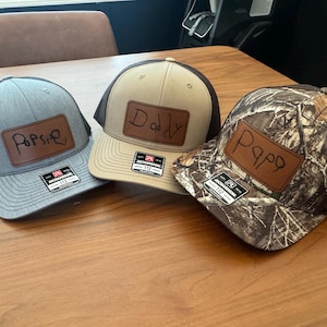 May include: Three trucker hats with personalized leather patches. One hat is gray with a black mesh back and the word "Popsie". Another is tan with a brown mesh back and the word "Daddy". The third is camouflage with a brown mesh back and the word "Papa".