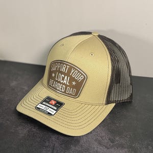 以下が含まれることがあります： メッシュバックのタンとブラウンのトラッカーハット。帽子の前面には「SUPPORT YOUR LOCAL BEARDED DAD」というテキストのパッチが付いています。帽子には湾曲したつばと調節可能なストラップが付いています。