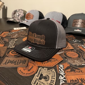 Może przedstawiać: Czarno-szara czapka z daszkiem z brązową skórzaną naszywką z napisem "Live Laugh Limp Bizkit". Czapka leży na stole z innymi naszywkami i naklejkami.