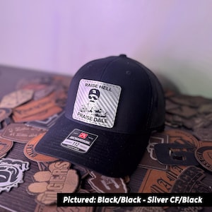 May include: A black trucker hat with a black mesh back. The hat has a silver carbon fiber patch with the text "Raise Hell" and "Praise Dale" and a cartoon image of a man with a mustache.