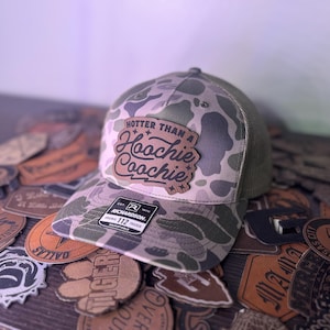 May include: A camouflage trucker hat with a brown leather patch that says "Hotter than a Hoochie Coochie" in a cursive font. The patch has small stars around the text. The hat is on top of a pile of leather patches.