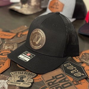 Peut inclure: Casquette de camionneur noire avec un patch en cuir marron qui dit "Uncle Danny's Grilled Cheese". Le patch a une image d'un sandwich au fromage grillé.