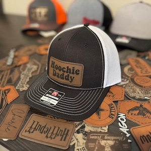 Op de afbeelding: Een zwarte en witte trucker-pet met een bruin leren patch waarop in een retro-lettertype "Hoochie Daddy" staat. De pet is omringd door verschillende leren patches met verschillende ontwerpen.