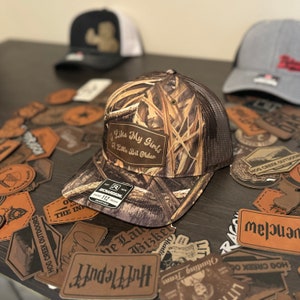 Puede incluir: Una gorra de camionero de camuflaje marrón y verde con una malla trasera marrón. La gorra tiene un parche de cuero marrón en la parte delantera que dice "Me gustan mis chicas un poco mayores".