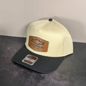Può includere: Cappellino da baseball beige e nero con una toppa in pelle con il logo Chevrolet e la scritta "CHEVROLET EST. 1911". Il cappellino ha una visiera curva e un bottone nero sulla parte superiore.
