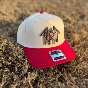 Puede incluir: Gorra de béisbol color crema con visera roja. La gorra presenta un diseño floral y el texto "OTTO" en una pegatina. La gorra tiene un diseño de perfil medio.