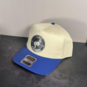 Peut inclure: Casquette de baseball Ford Bronco beige et bleu. La casquette a une visière bleue et une couronne beige. L'avant affiche un emblème circulaire avec "FORD BRONCO" et un graphique de cheval. Coupe classique et profil moyen.