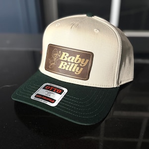 Puede incluir: Una gorra de béisbol blanca y verde con un parche de cuero marrón que dice "Baby Billy" en letras doradas. La gorra tiene un ajuste clásico y un perfil medio.