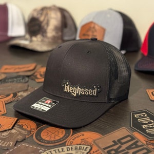 May include: Black trucker hat with a black mesh back and a gold embroidered patch that says "bleghssed".