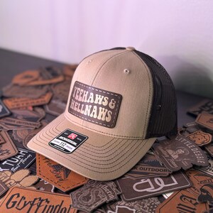 Puede incluir: Una gorra de camionero de color beige y marrón con un parche de cuero marrón que dice "YEEHAWS & HELLNAWS" en letras doradas. La gorra está sobre una pila de parches de cuero marrón.