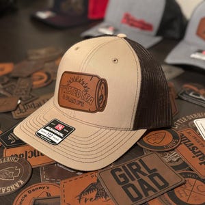 May include: A brown and tan trucker hat with a brown leather patch that says "Twisted Tea & Plan B's". The hat is surrounded by various leather patches with different designs and text.