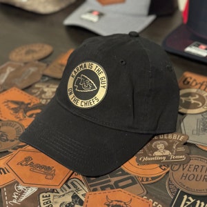 Puede incluir: Una gorra de béisbol negra con un parche de cuero dorado que dice "Karma is the guy on the Chiefs". El parche presenta un logotipo rojo y blanco de los Kansas City Chiefs.