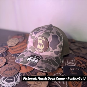 May include: A camouflage baseball cap with a brown leather patch featuring the state of Georgia and the text "EST. 1785." The cap has a mesh back and a curved brim. The text "Pictured: Marsh Duck Camo - Rustic/Gold" is at the bottom.