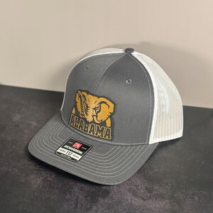 Könnte beinhalten: Grau-weiße Trucker-Cap mit dem Wort "ALABAMA" und einem goldenen Elefantenlogo auf der Vorderseite. Die Kappe hat einen gebogenen Schirm und einen verstellbaren Druckknopfverschluss.