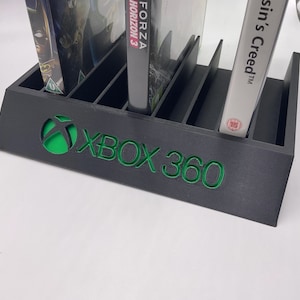 Puede incluir: Soporte de plástico negro con ranuras para guardar videojuegos. El soporte tiene un logotipo de Xbox 360 verde y contiene varias cajas de videojuegos, incluyendo una para Forza Horizon 3 y otra para Assassin's Creed.