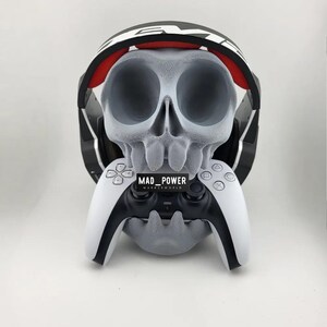 以下が含まれることがあります： グレーの頭蓋骨型ゲームヘッドセットとコントローラーのスタンド。 赤いアクセントが付いた白と黒のヘッドセットと、白と黒のコントローラーを保持します。 コントローラーには「MAD_POWER」の文字が見えます。