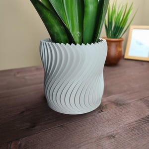 May include: A gray, textured, wavy ceramic planter with a green plant inside. The planter is sitting on a wooden surface.