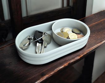 May include: A grey oval-shaped tray with a smaller round dish inside. The tray has a raised edge and the dish has a lip. The tray holds keys and the dish holds coins.