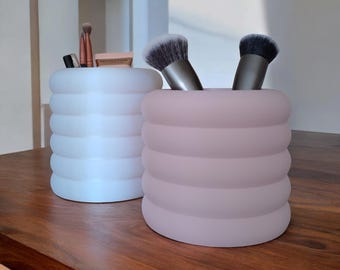 May include: Two cylindrical makeup brush holders, one white and one light pink, with a wavy, textured design. The holders are filled with makeup brushes.