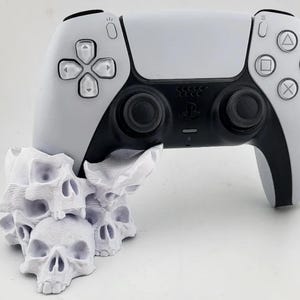 May include: A white PlayStation 5 DualSense controller is resting on a white skull-shaped stand. The controller has black accents and buttons.