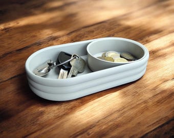 May include: A grey oval-shaped tray with a smaller circular tray inside. The larger tray has a raised edge and the smaller tray has coins inside. Keys are in the larger tray.