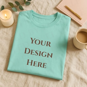 May include: A light teal t-shirt with the words "YOUR DESIGN HERE" printed in brown. The shirt is folded neatly on a beige surface, with a lit candle, a cup of coffee, a pen, and a notebook nearby.