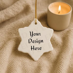 May include: A white star-shaped ornament with the text "Your Design Here" in black lettering, suspended by a gold string. A lit candle in a beige ceramic holder sits nearby, all on a textured beige surface.