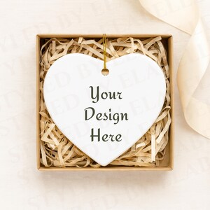 Könnte beinhalten: Ein weißer herzförmiger Ornament mit dem Text "Your Design Here" in einer Geschenkbox, gefüllt mit geschreddertem Papier. Der Ornament hat eine goldene Schnur zum Aufhängen. Ein cremefarbenes Band befindet sich im Hintergrund.