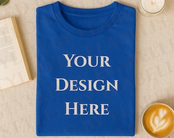 Op de afbeelding: Een koningsblauw T-shirt met de witte tekst "YOUR DESIGN HERE". Het shirt is op een neutrale ondergrond gevouwen, samen met een boek, een brandende kaars en een kopje koffie. Het T-shirt is klaar voor een aangepast ontwerp.