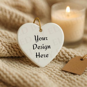 May include: A white heart-shaped ornament with the text "Your Design Here" in black. The ornament hangs from a gold string. A lit candle and a brown tag are in the background, all on a beige knitted surface.