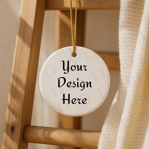 Può includere: Un ornamento rotondo in ceramica bianca con la scritta "Your Design Here" in corsivo nero. L'ornamento è appeso a un filo dorato, con una scala di legno e una coperta color crema sullo sfondo.