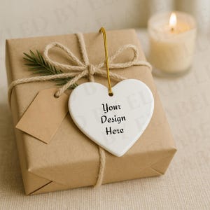 May include: A brown paper-wrapped gift tied with twine and a sprig of greenery. A white heart-shaped ornament with the text "Your Design Here" hangs from a gold string. A small brown tag is attached.