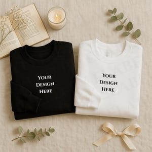Könnte beinhalten: Ein schwarzes und ein weißes Sweatshirt, jeweils mit dem Text "YOUR DESIGN HERE". Die Sweatshirts werden auf einer neutralen Oberfläche präsentiert, zusammen mit einer brennenden Kerze, einem offenen Buch und dekorativen Zweigen.