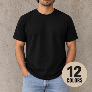 Puede incluir: Una camiseta negra lisa de manga corta con cuello redondo. La prenda se lleva con unos vaqueros de mezclilla azul claro. Un gráfico circular de color beige en la esquina inferior derecha muestra el texto negro 12 COLORS, indicando opciones.