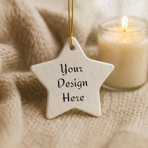 May include: A white star-shaped ornament with the text "Your Design Here" in black, hanging by a gold string. A lit candle in a glass jar is in the background, set against a beige, textured fabric.
