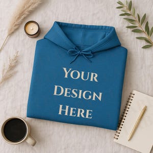 May include: A teal blue hoodie with the text "YOUR DESIGN HERE" in white. The hoodie has a drawstring and a hood. A cup of coffee, a candle, a notebook, and a pen are also in the image.