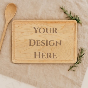 May include: A light-colored wooden cutting board with a rectangular shape and a recessed border. The text "YOUR DESIGN HERE" is engraved on the board. A wooden spoon and sprigs of rosemary are placed around the board.