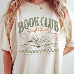 May include: An off-white t-shirt featuring a graphic with the text BOOK CLUB, Fresh Chapters, PAGE-TURNER, and SHELF FRESH. The design includes an open book illustration, leafy branches, a checkered pattern, and the words NOVELS, PROSE, POETRY.