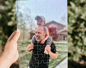 Könnte beinhalten: Ein rechteckiges Puzzle mit einem Foto eines Mannes, der ein Kind auf seinen Schultern trägt. Das Bild zeigt den lächelnden Mann und das Kind mit einer Mütze. Das Puzzle wird vor einem verschwommenen grünen Hintergrund gehalten.