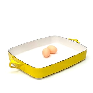 May include: A rectangular yellow baking dish with a white interior and two eggs. The dish has two yellow handles and a black trim around the rim. The eggs are light brown.