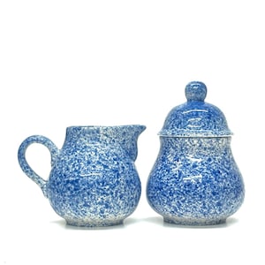 May include: A blue and white speckled ceramic creamer and lidded sugar bowl set. The creamer has a handle and spout. The sugar bowl has a rounded shape with a lid and a small knob on top. The set is on a white background.