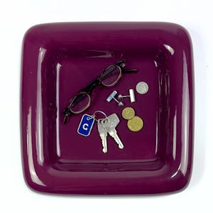 May include: A square, deep purple ceramic catch-all tray with rounded corners. Inside the tray are a pair of dark-rimmed eyeglasses, a set of silver-toned cufflinks, two metal coins, and a keyring with two keys and a blue tag featuring the letter C.